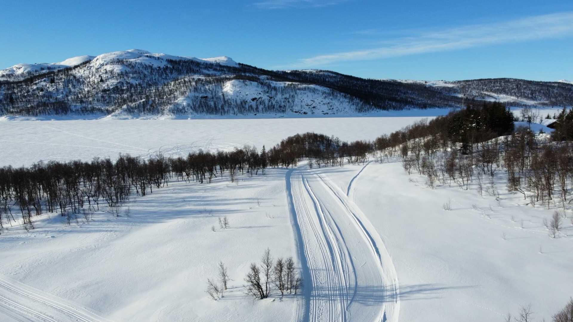Vinterlandskap med skiløyper og fjell bak en snødekt innsjø under blå himmel.