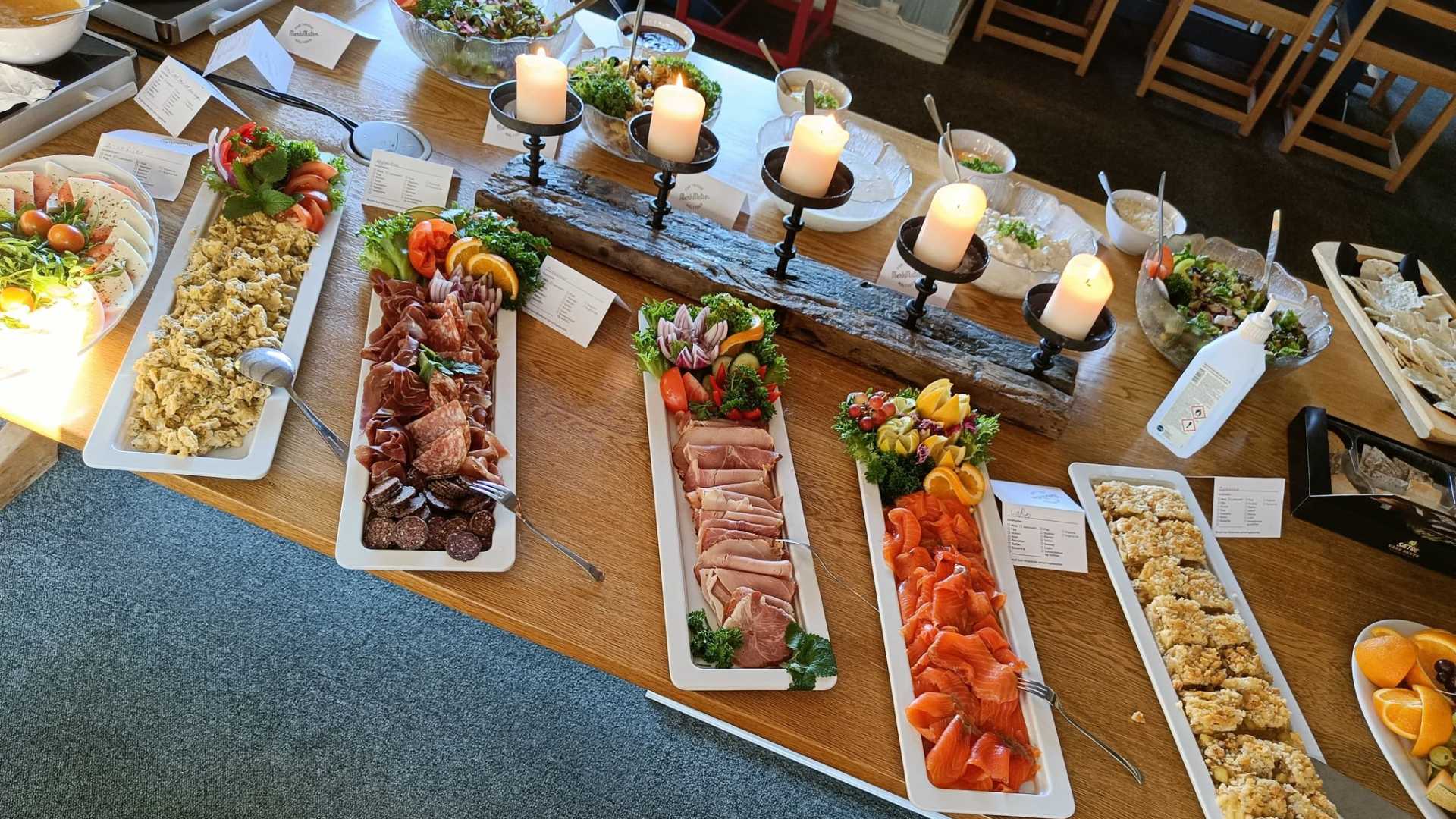 Buffet med diverse delikatesser: kjøttpålegg, røkt laks, salater og osteanretninger, dekorert med lys og grønt.