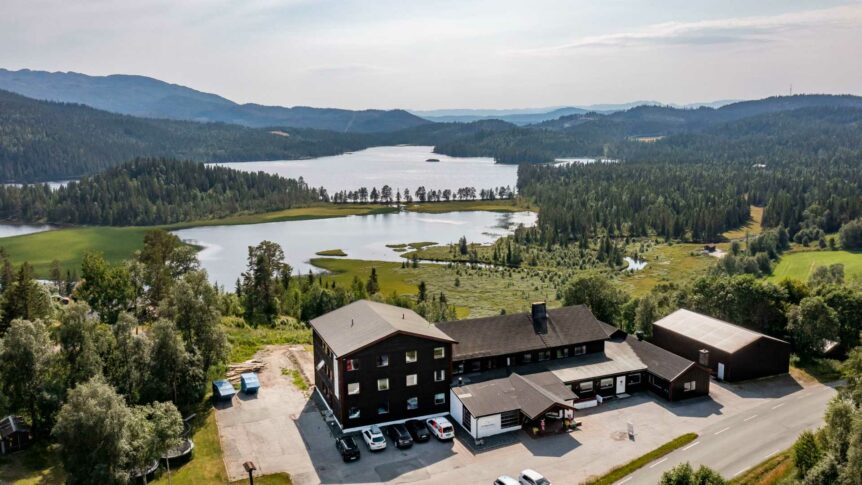 Flyfoto av et trehus nær en innsjø i frodig norsk landskap med fjell og skog i bakgrunnen.