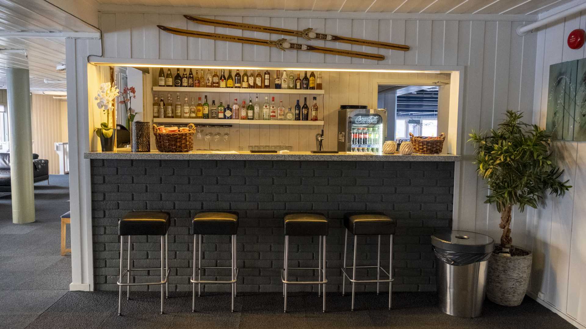 Koselig bar med ski som dekorasjon, fire barkrakker og variert utvalg av flasker på hyller i bakgrunnen.