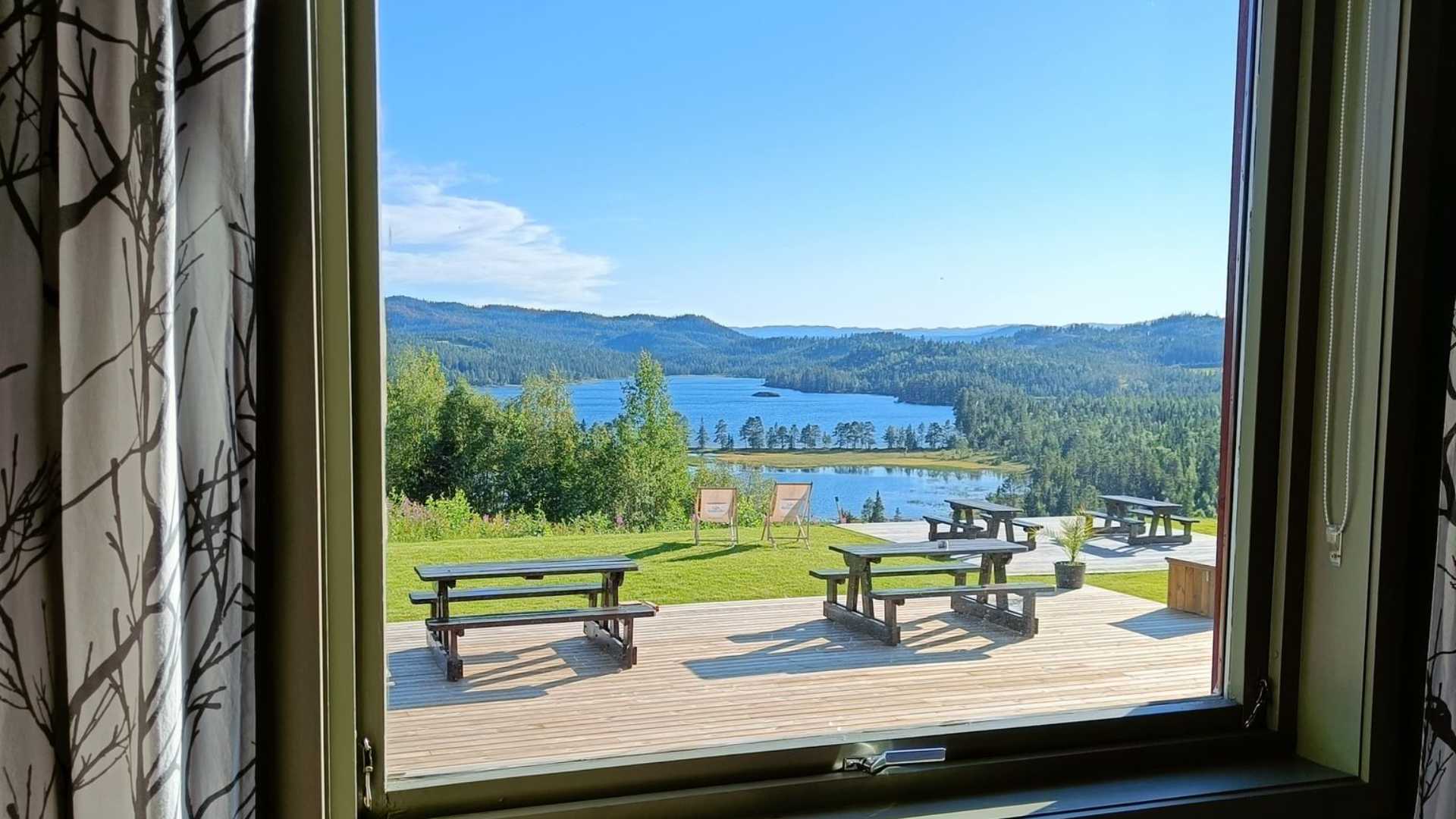 Utsikt fra vindu mot innsjø og skogkledde åser, med terrasse og benker i forgrunnen. Sommerdag i norsk natur.