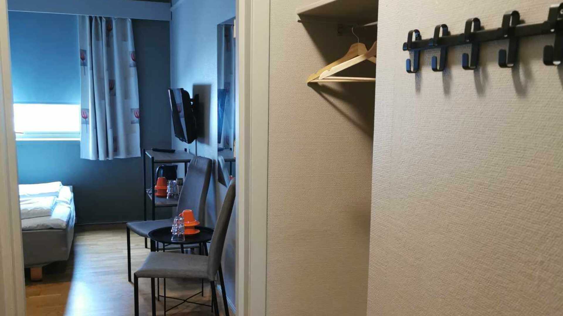 Hotelrommet har seng, bord med stoler, og TV ved vinduet. En åpen garderobe og kleshengere sees ved inngangen.