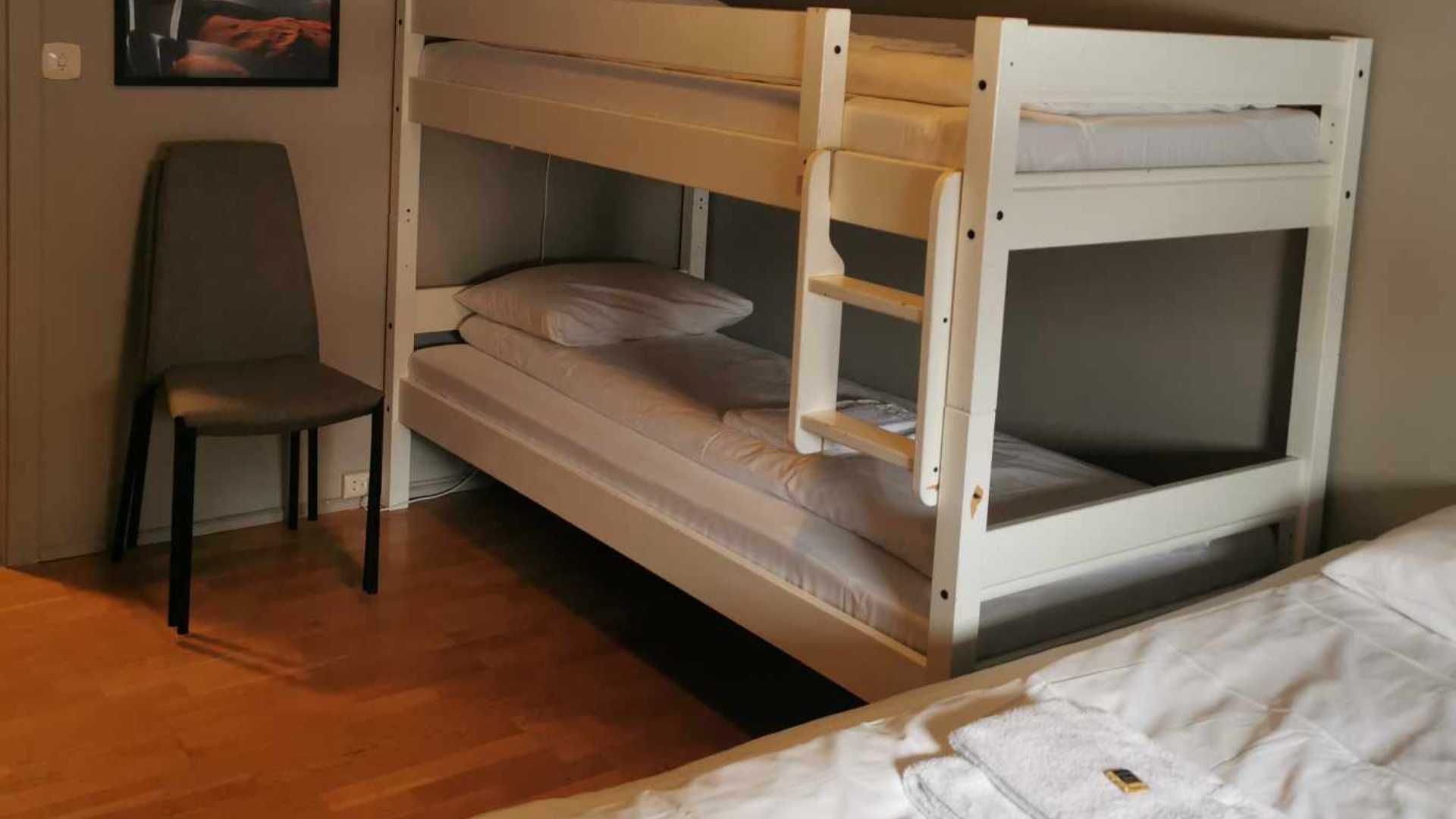 Køyeseng med hvitt sengetøy ved siden av stol på soverom med tregulv og veggkunst.