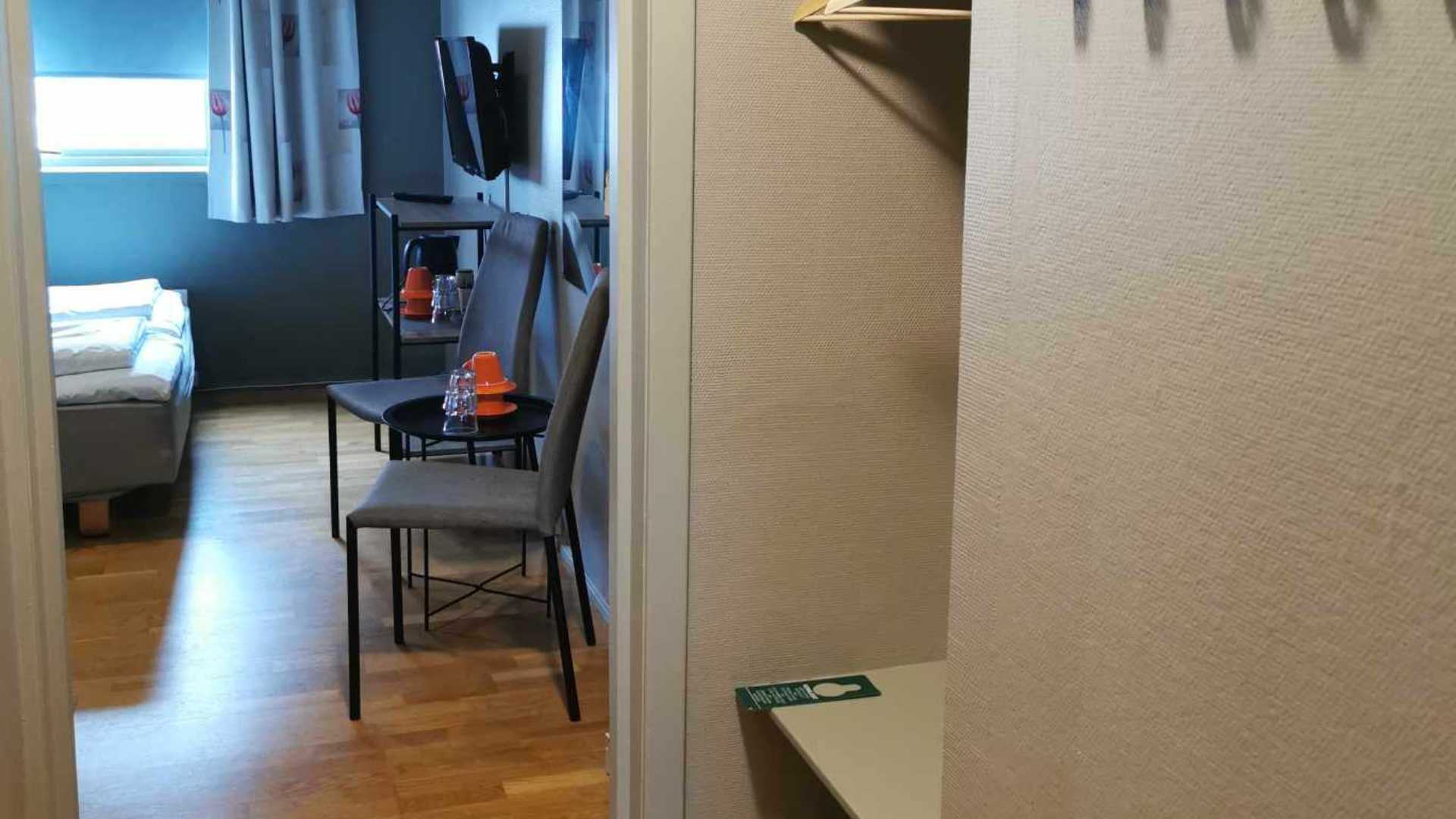 Hotellrom med seng, TV, stoler og bord på tregulv, sett fra inngangen. Minimalistisk interiør i nøytrale farger.
