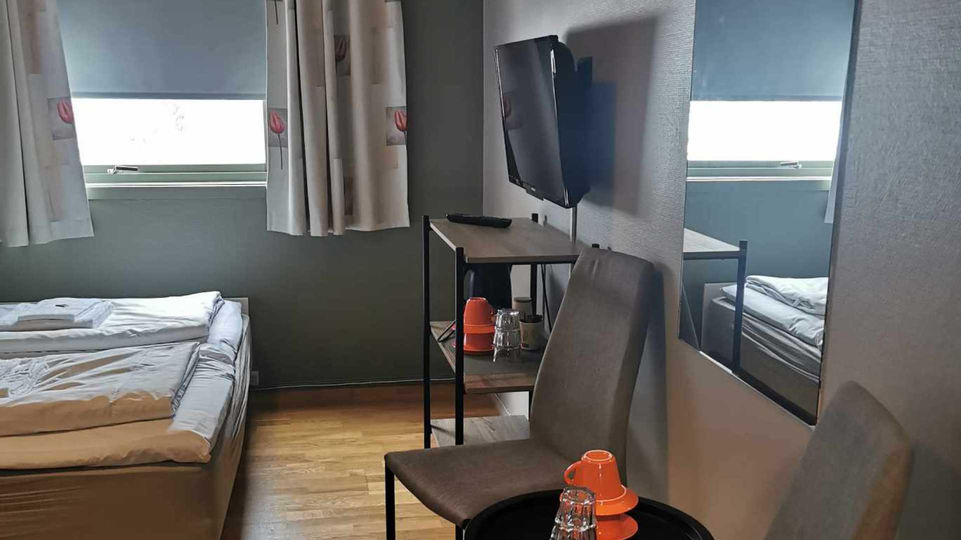 Moderne hotellrom med seng, TV og te-sett på bord. Lyse gardiner og tremøbler gir en koselig atmosfære.