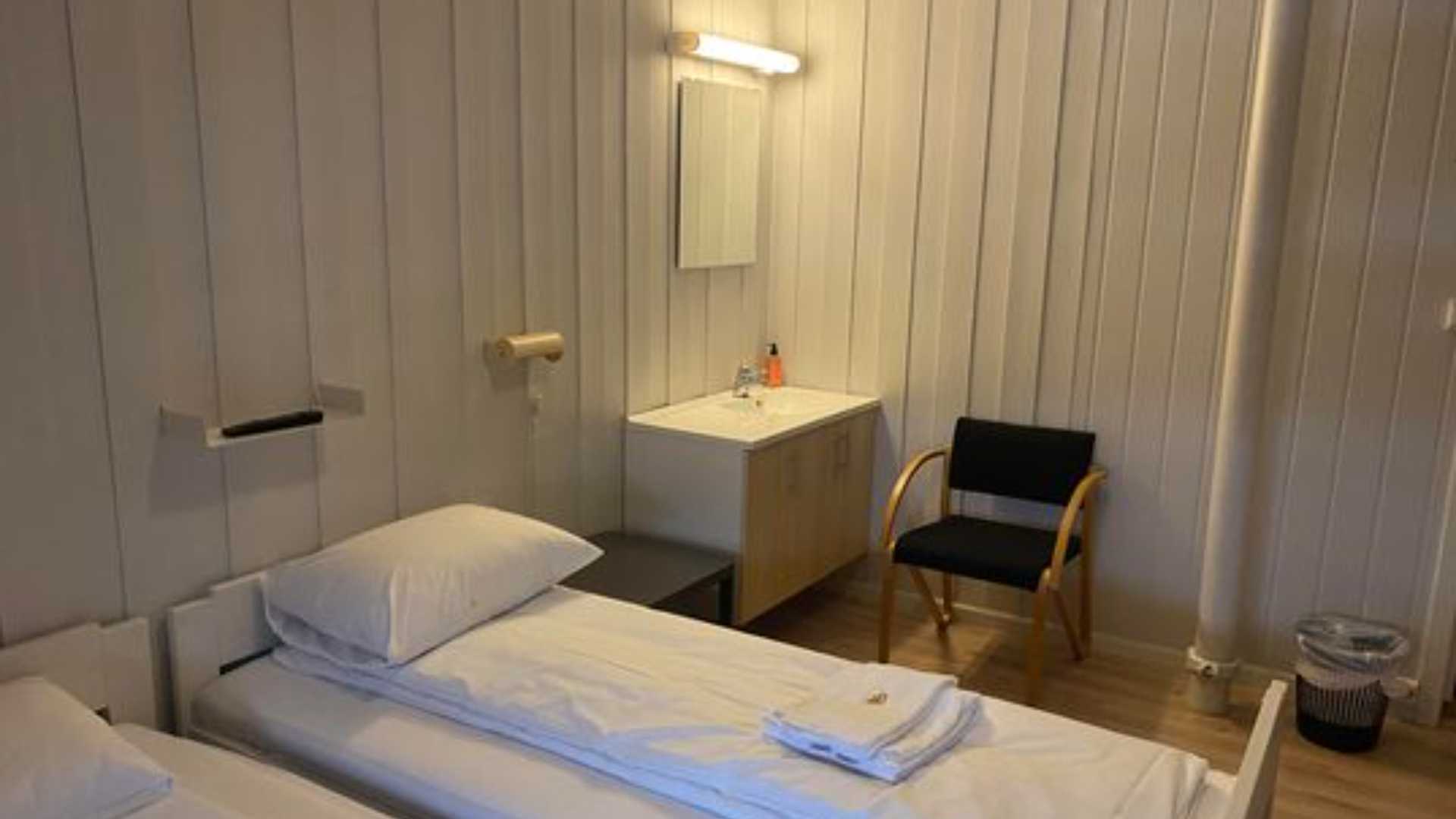 Enkel og koselig rom med to senger, hvite vegger, speilbord og stol i hjørnet.