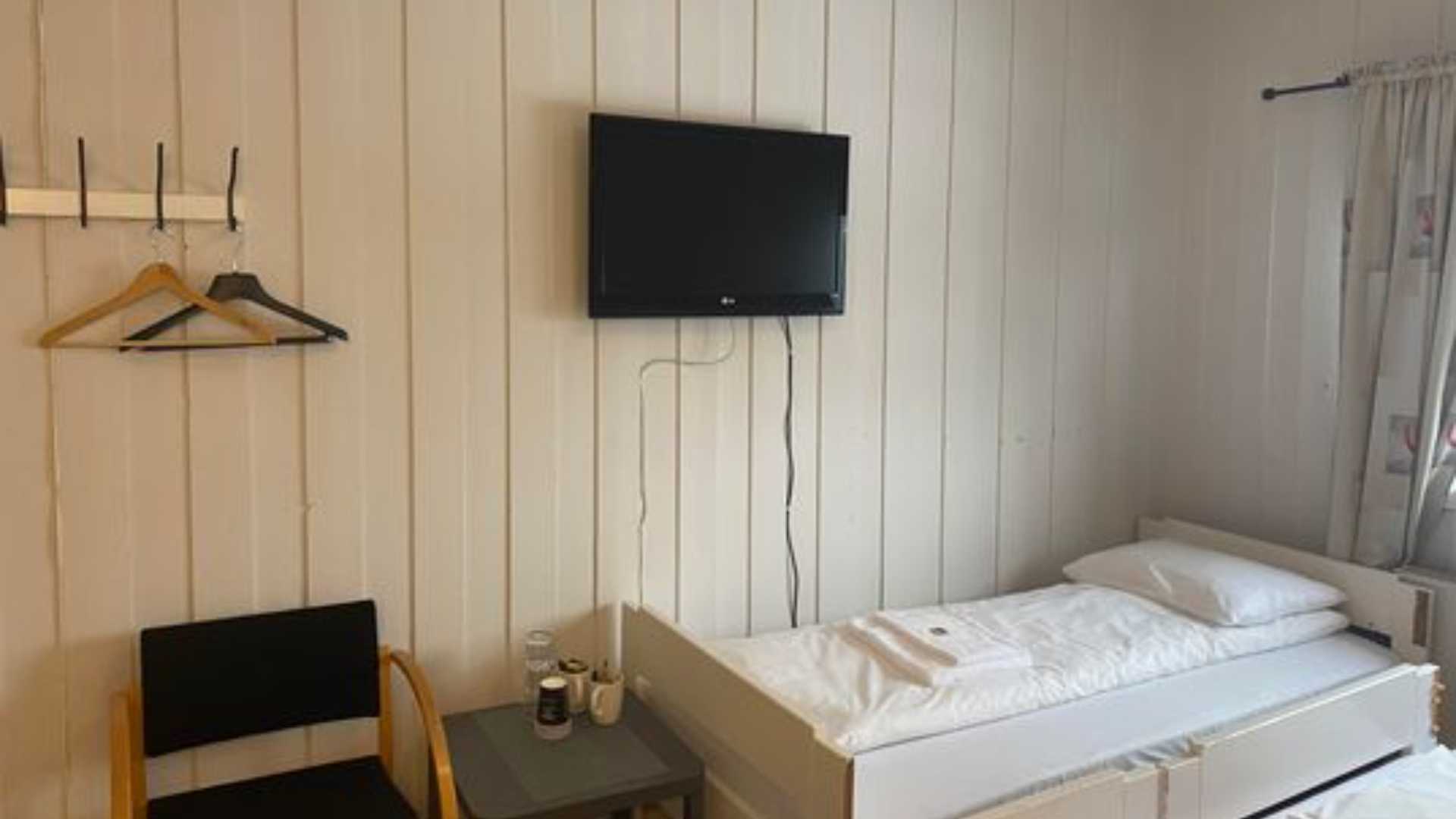 Enkel hotellrom med seng, TV, stol og kleshenger. Lyse vegger og minimalistisk interiør.