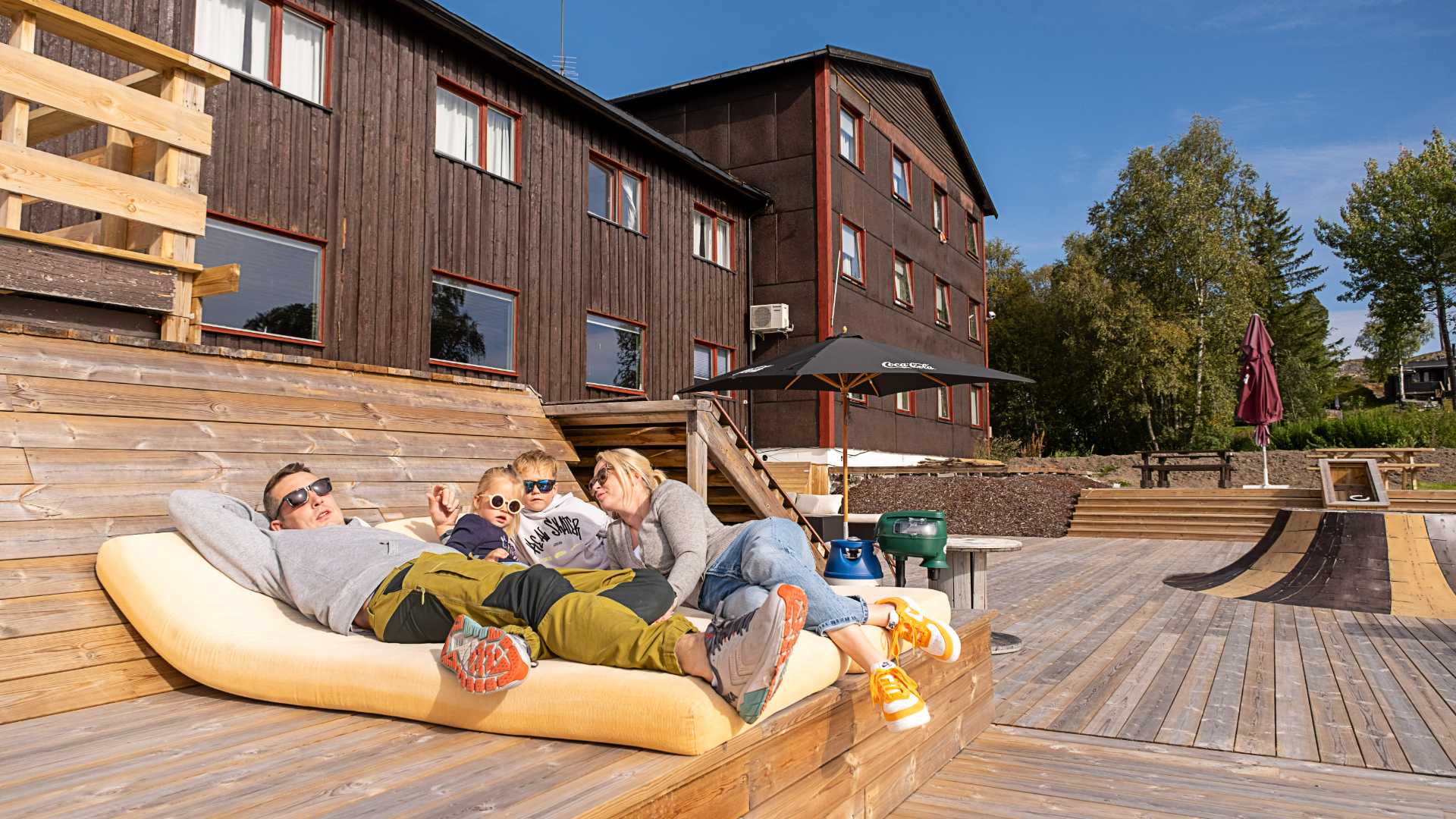 Familie slapper av i solskinnet på terrasse ved trehus, sommerstemning.
