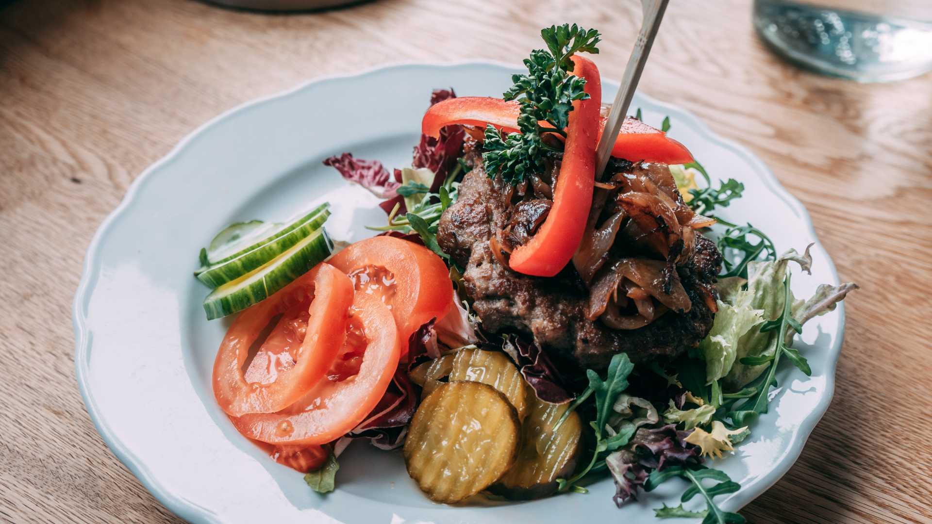 Tallerken med saftig hamburger, tomat, agurk og syltetøy, servert med løk og salat på et rustikt trebord.