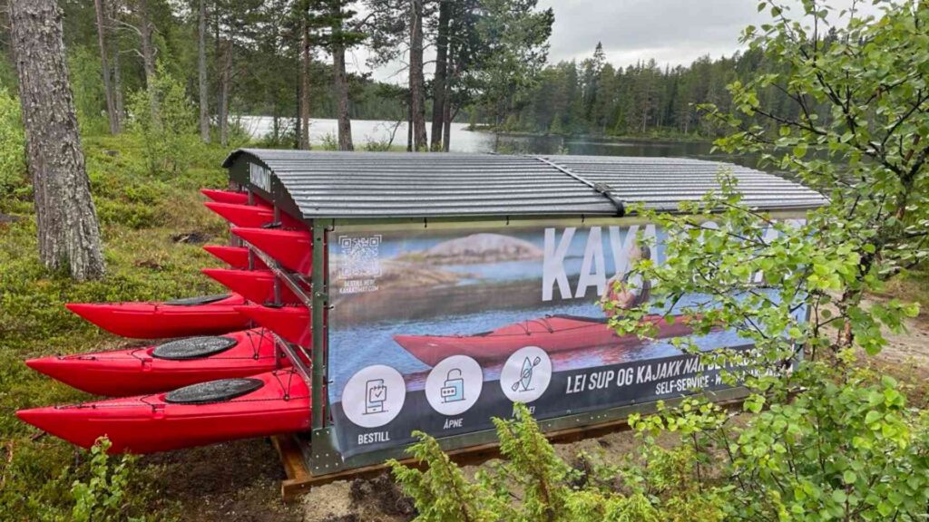 Røde kajakker på rad utenfor et kajakkutleie ved innsjøen, omkranset av skog.