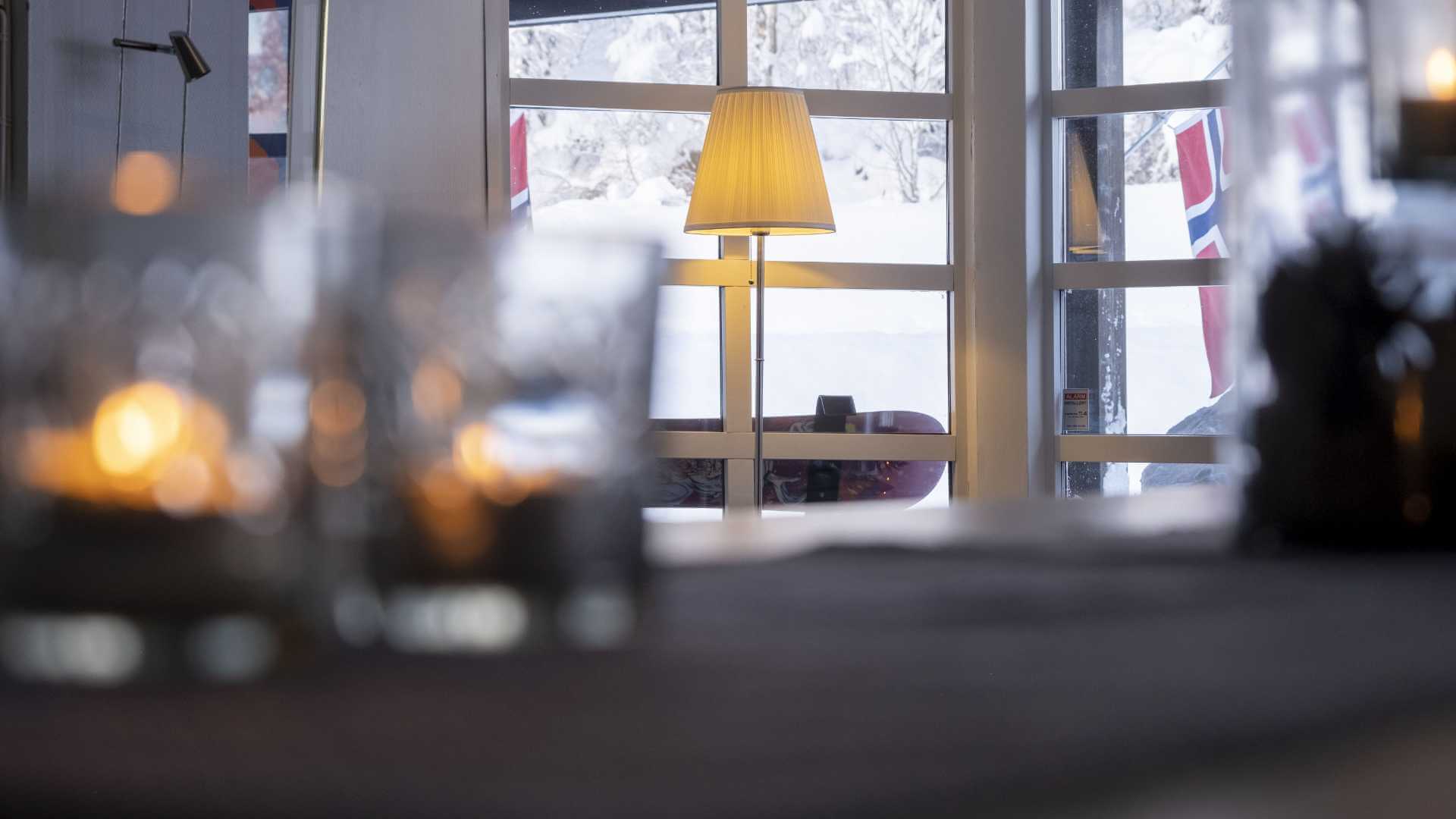 Koselig interiør med tente lys og lampe, vinterlandskap utenfor med norsk flagg i vinduet.