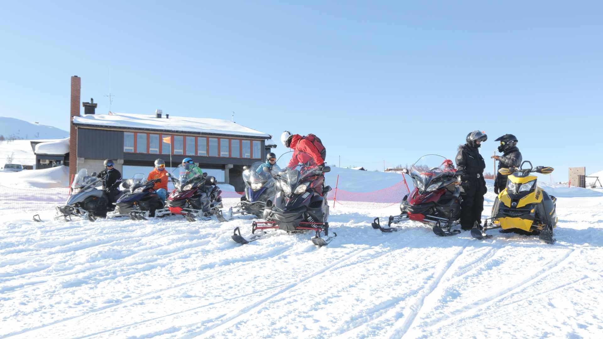 Personer på snøscootere foran en fjellhytte i solfylt vintervær.