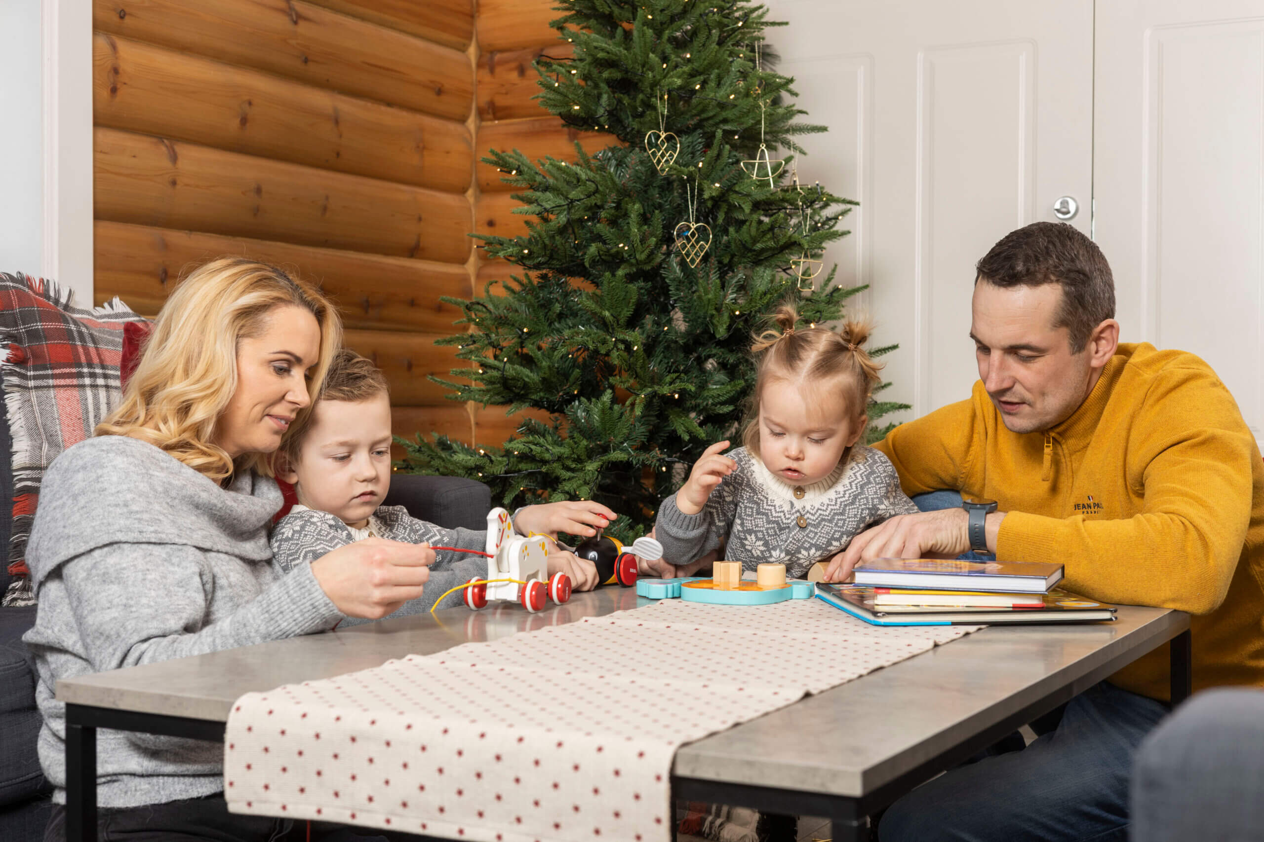 Familie ved juletre, leker med barna ved bordet, koselig julefeiring i stua.
