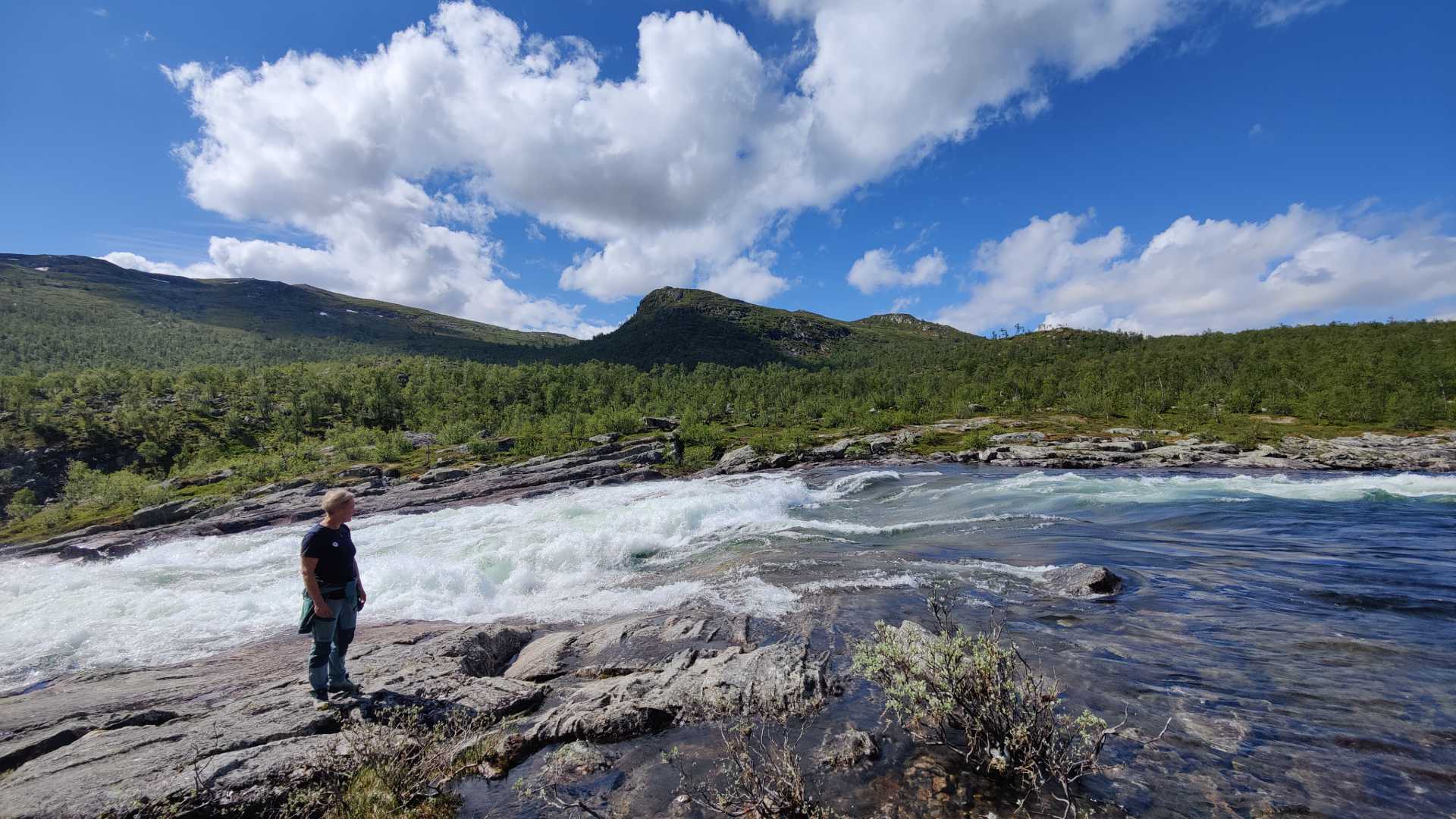 Ukjent person ved fjellbekk omringet av grønn skog og fjell under en solrik himmel med skyer.