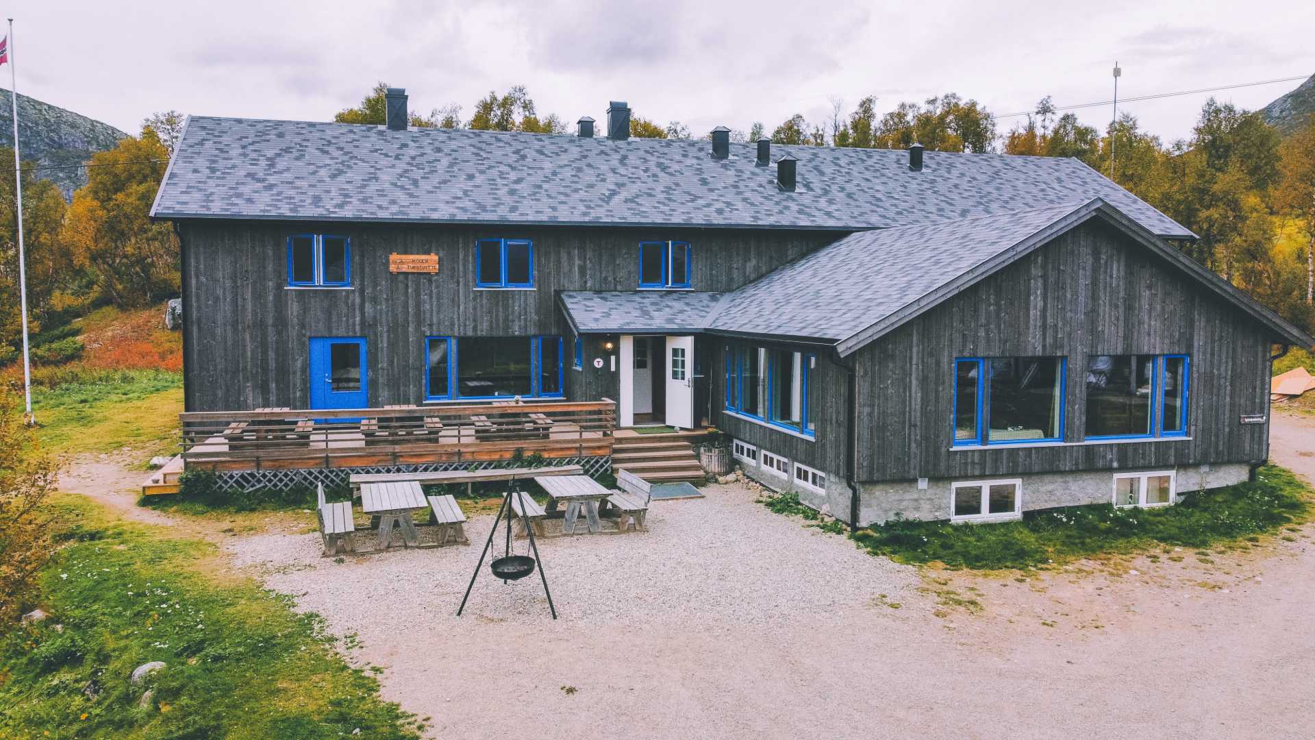 Stor hytte i tre omgitt av natur med blå dører og vinduer, beige tak og sitteplasser utenfor.