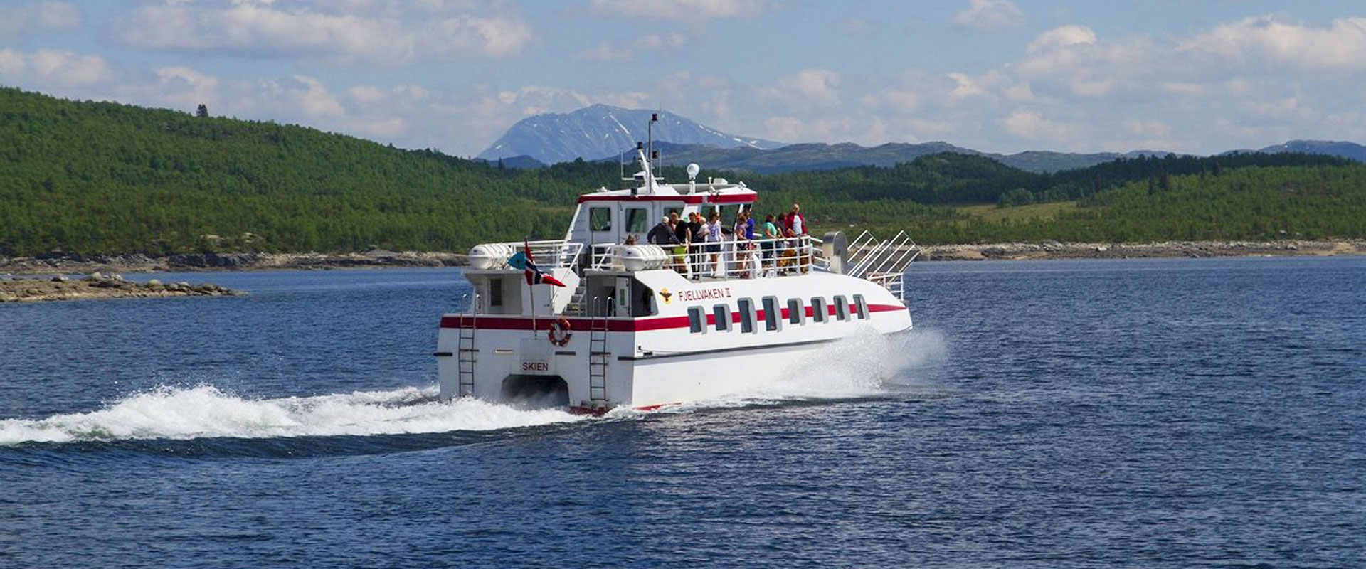 En hvit passasjerbåt på et norsk fjord med grønne åser og fjell i bakgrunnen.