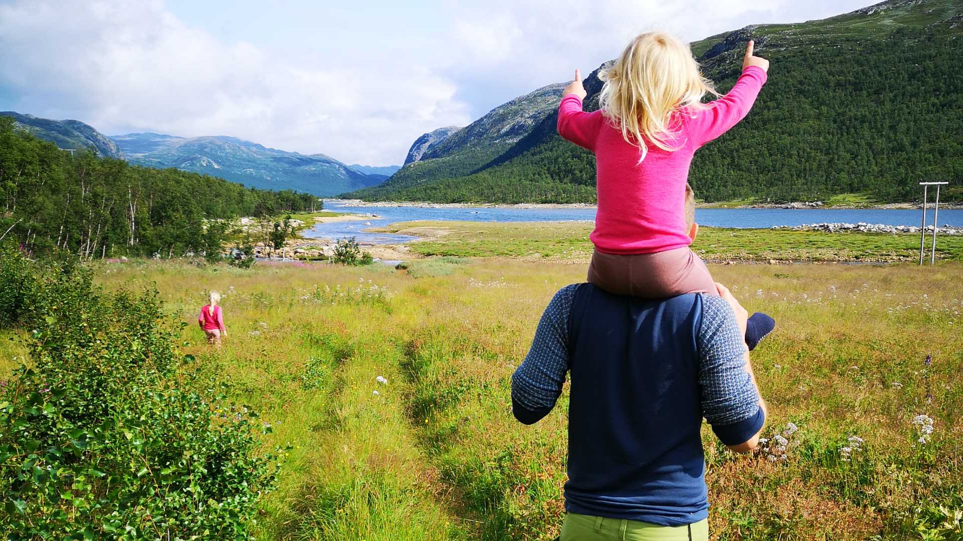 Far og datter ute i norsk natur ved innsjø, med fjell i bakgrunnen. Jente på skuldrene peker mot fjellet.