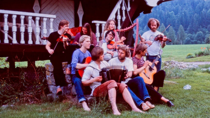 Gruppemusikere spiller tradisjonelle instrumenter utendørs foran en trebygning i naturskjønne omgivelser.