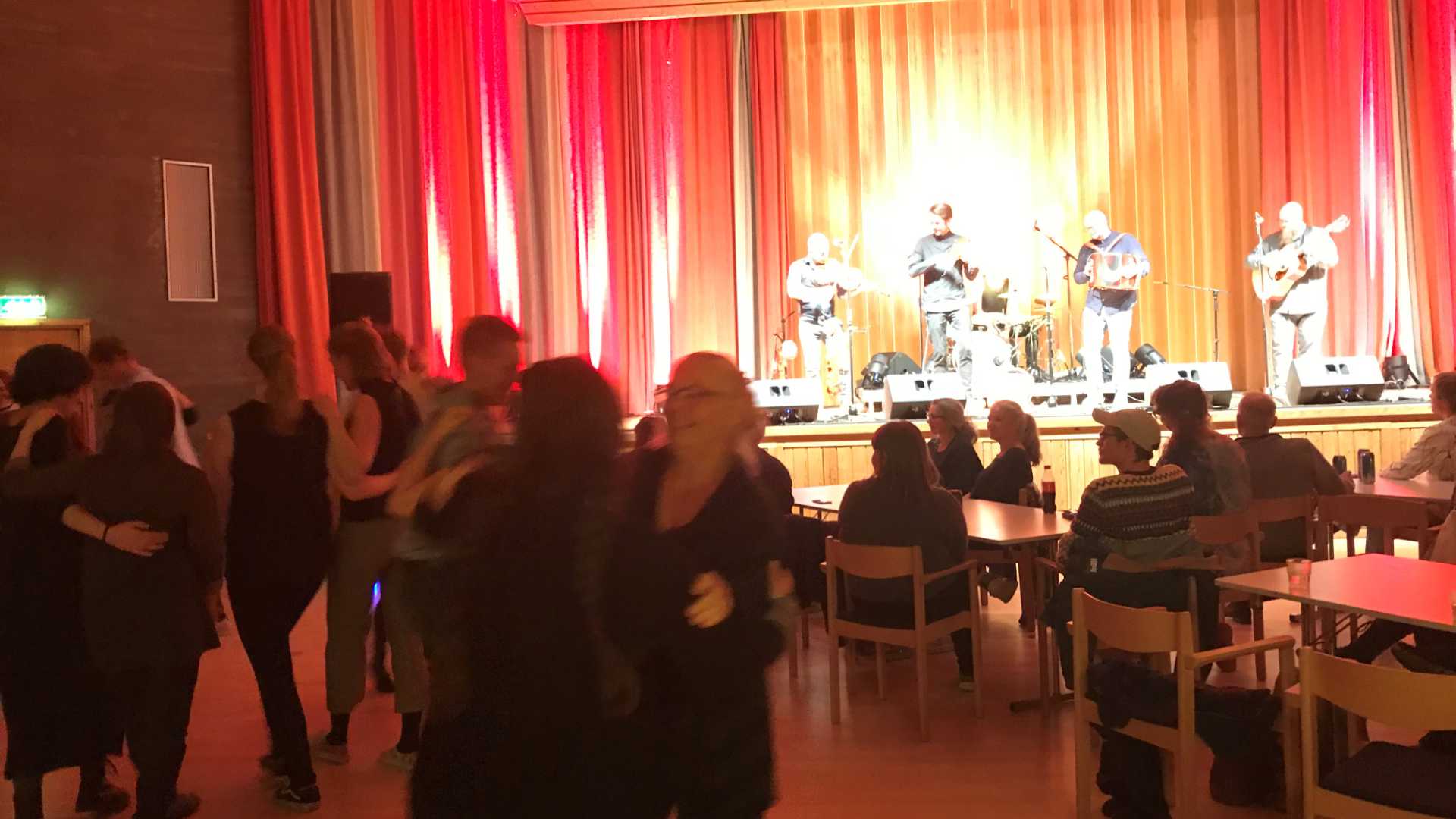 Dansende par og bandoppvisning med livemusikk på en scene i et fargerikt lokale.