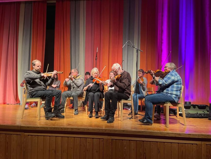 En gruppe musikere spiller fiolin på en scene med fargerik bakgrunnsbelysning.