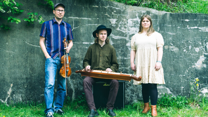Tre musikere med instrumenter foran en grå murvegg, natur rundt dem.