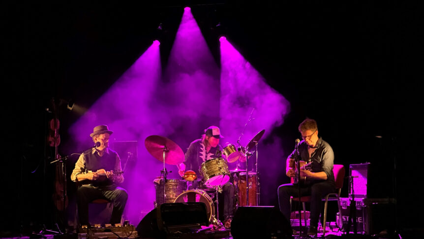 Tre musikere spiller under lilla scenelys på en konsert med gitar og trommesett.
