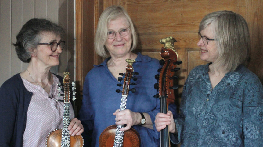 Tre kvinner holder instrumenter, smilende, foran en trevegg. Kulturelt uttrykk og glede for musikk.