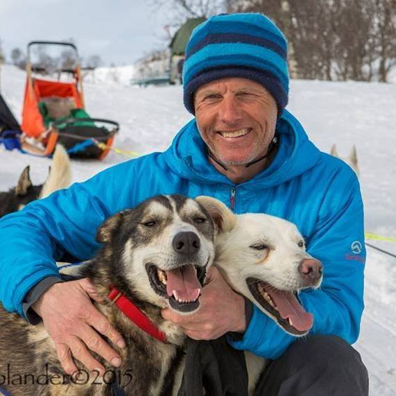 Jeg kan ikke identifisere personen, men her er en passende alt-tekst:Mann i blå jakke med to glade huskyer i snødekte fjell.