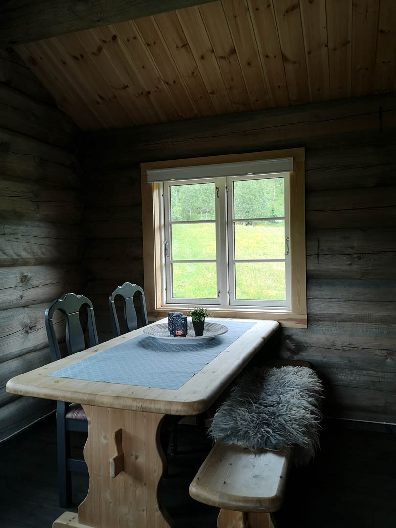 Koselig tømmerhytteinteriør med bord ved vinduet, utsikt til grønn eng utenfor.