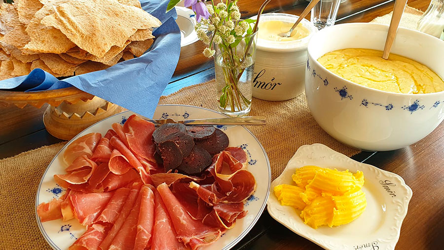 Tradisjonell norsk frokost med spekemat, flatbrød, smør og rømme i koselig borddekor med blomster.