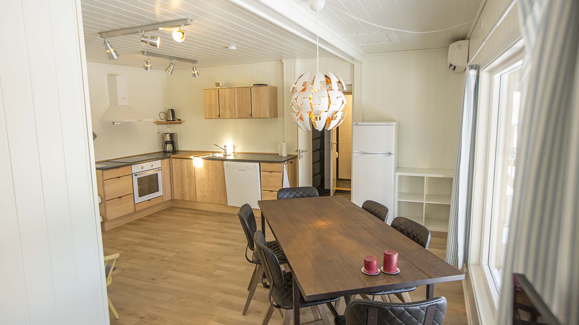 Moderne kjøkken med spisebord, trestoler, hvitevarer og lyse farger. Store vinduer gir naturlig lys og romfølelse.