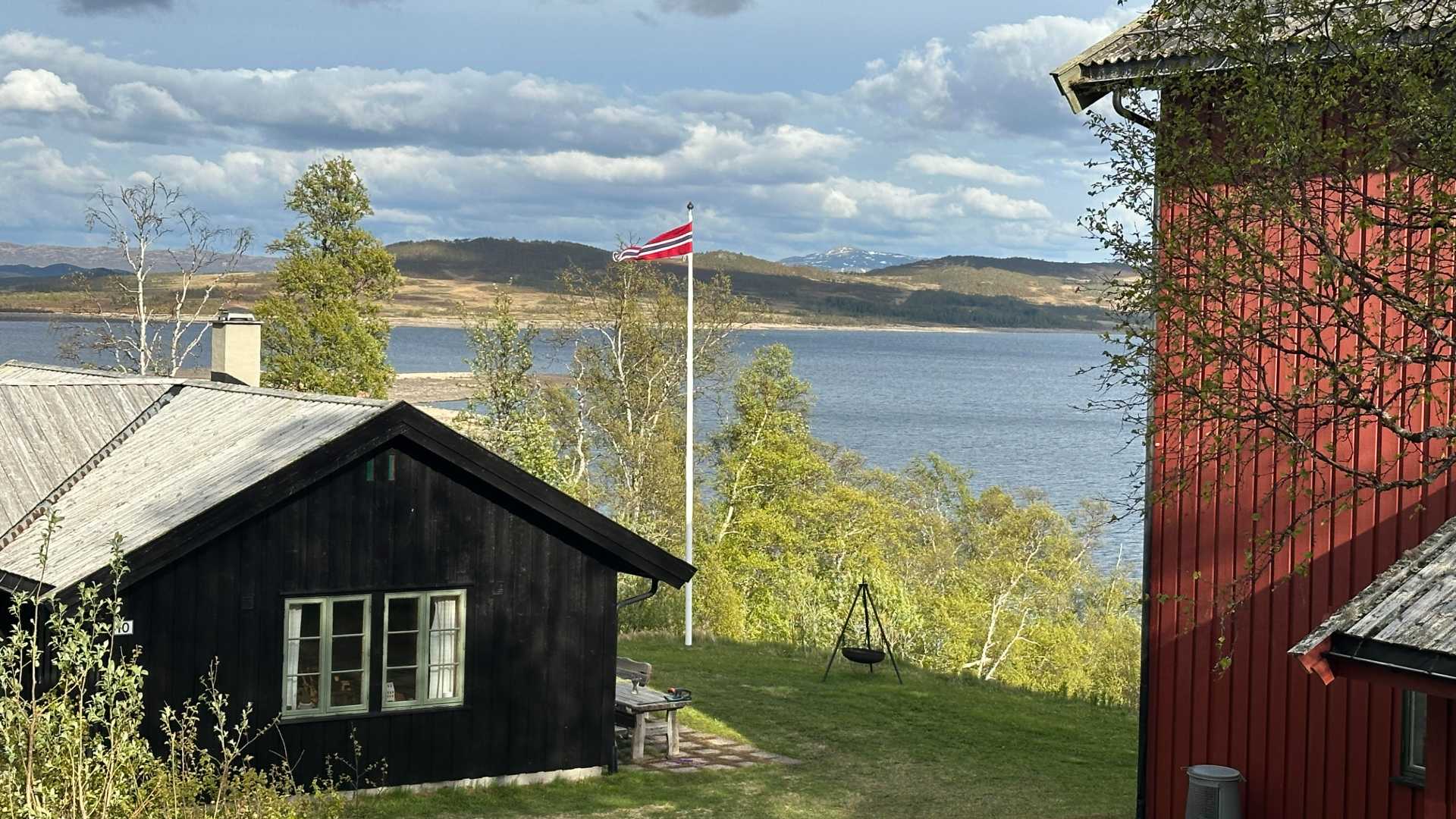 Norsk landskap med hytter ved innsjø, flagg på stang, trær og fjell i bakgrunnen under en skyet himmel.