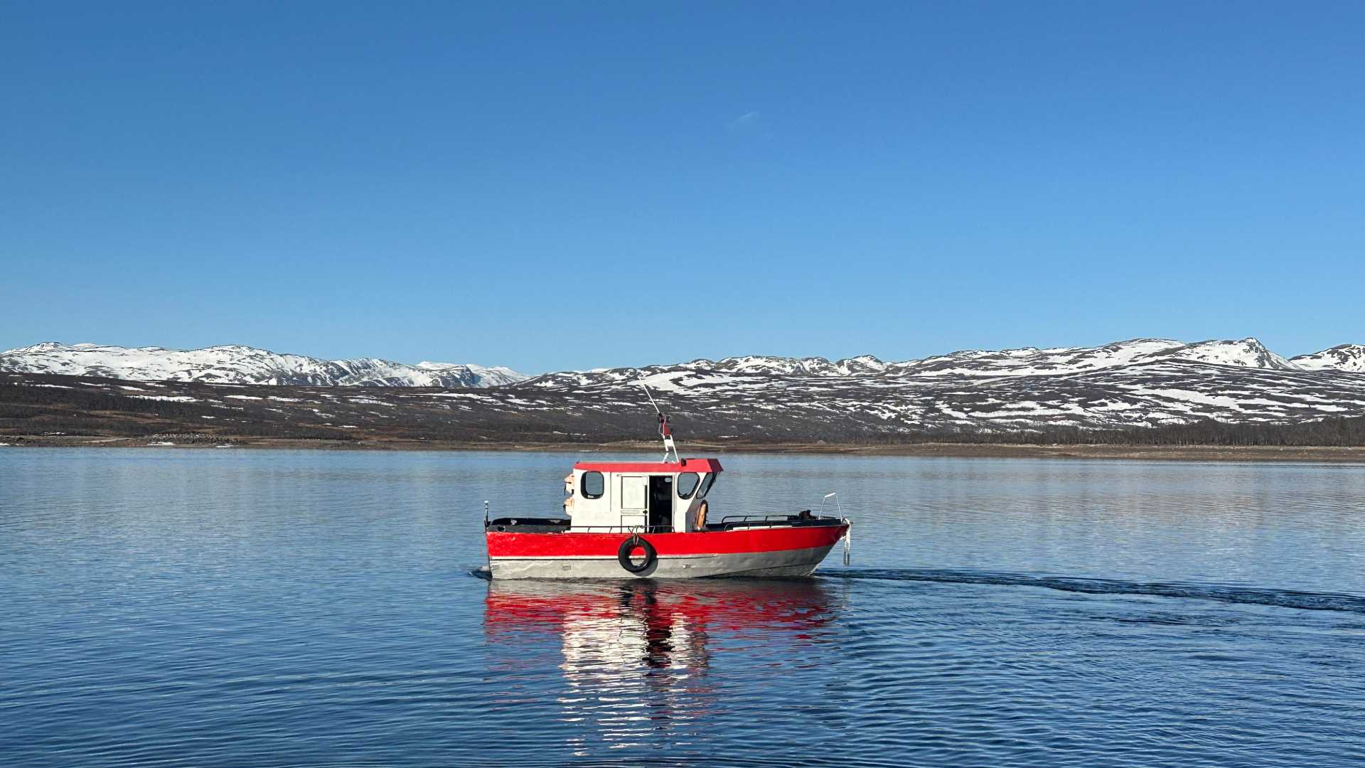 Rød og hvit båt på rolig innsjø med snødekte fjell i bakgrunnen under en klar blå himmel i Norge.