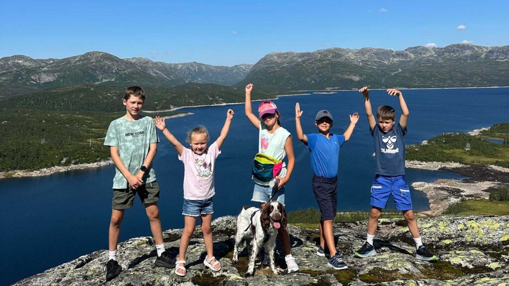 Barn og hund på fjelltur med utsikt over innsjø og fjell i bakgrunnen under blå himmel.
