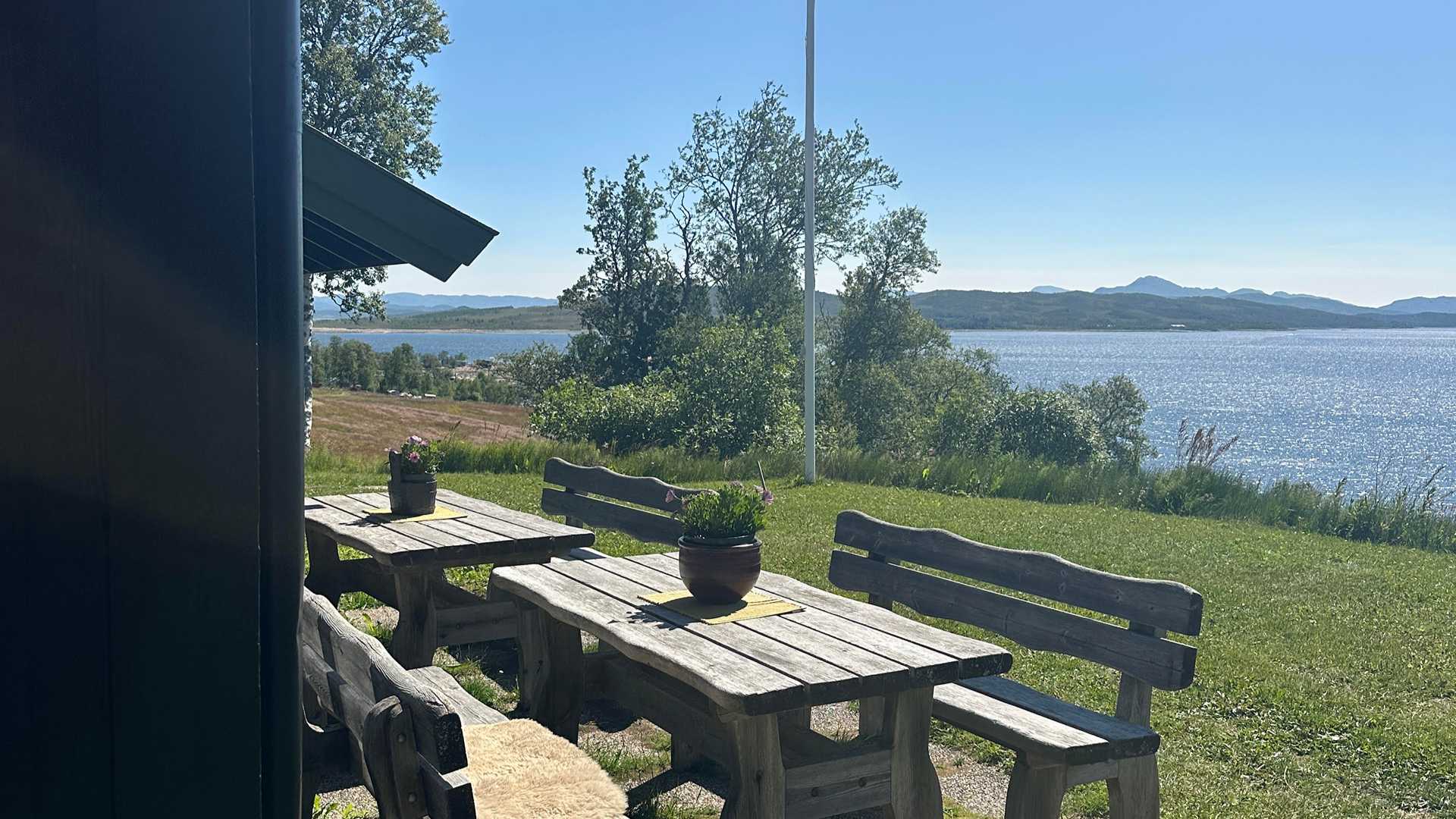 Trebord med utsikt over innsjø og fjell, omgitt av grønt gress og blå himmel.