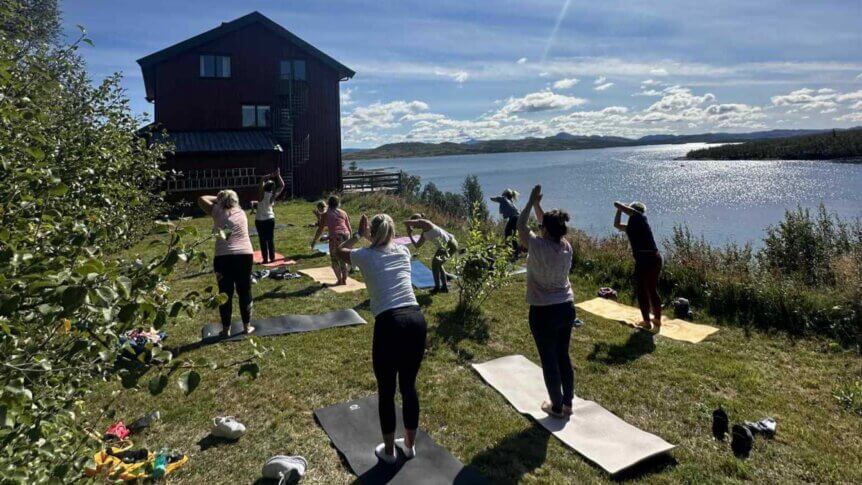 En gruppe mennesker gjør yoga utendørs ved en innsjø, med en rød bygning i bakgrunnen.