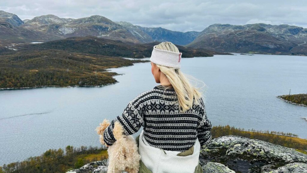 Kvinne i ullgenser med hund nyter utsikten over en norsk innsjø og fjellandskap.
