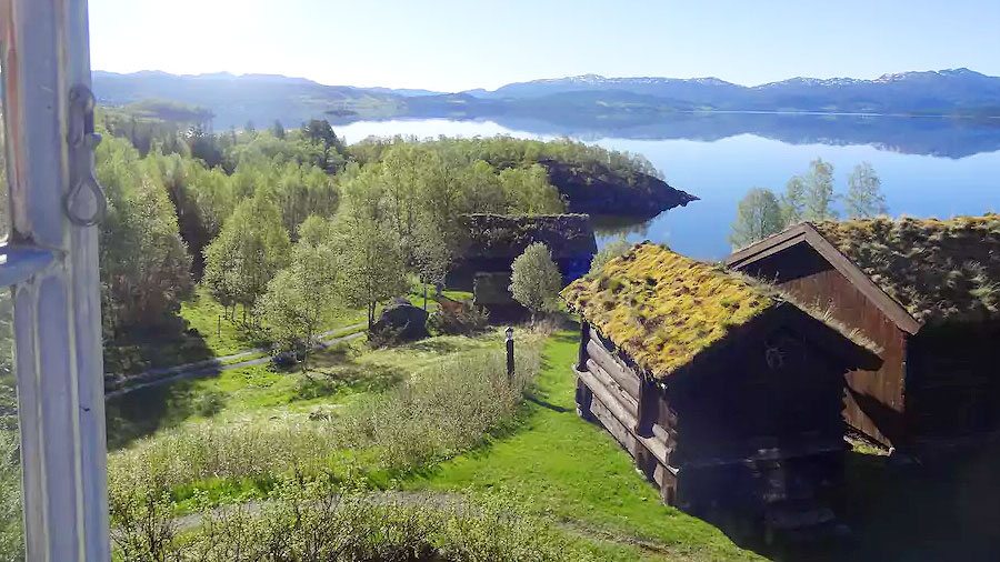 Gressdekte hytter ved en klar innsjø omringet av fjell og skog i Norge.