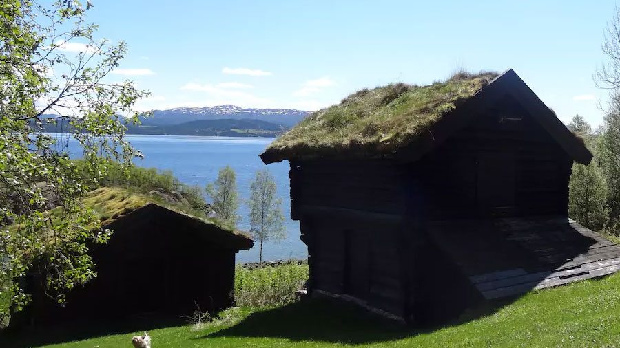 To gamle trehytter med torvtak ved en innsjø, omgitt av grønt gress og trær, med snødekte fjell i bakgrunnen.