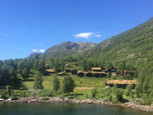 Gressdekte hus i en fredelig norsk dal, omgitt av skog og fjell under en blå himmel.