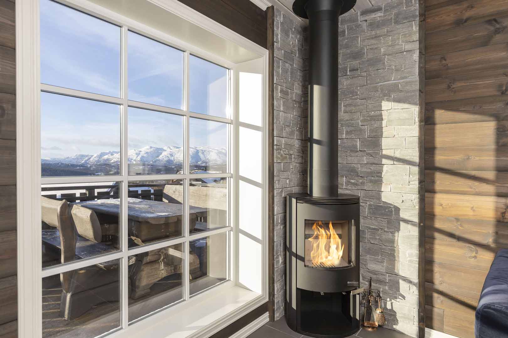 Peis i koselig hytteinteriør med utsikt over snødekte fjell gjennom stort vindu. Perfekt for vinterkvelder.