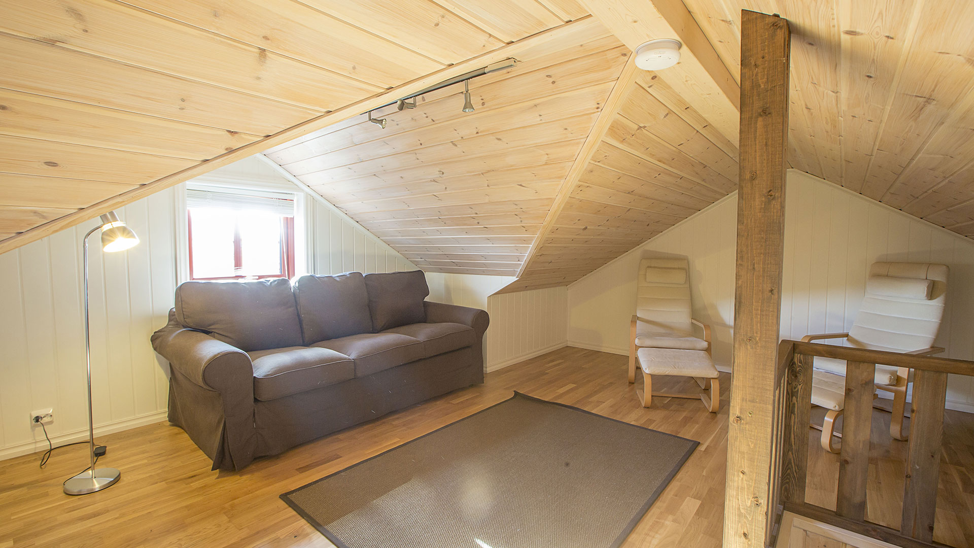 Lyst loftrom med brun sofa, to stoler, og stålampe under skråtak av trepanel.