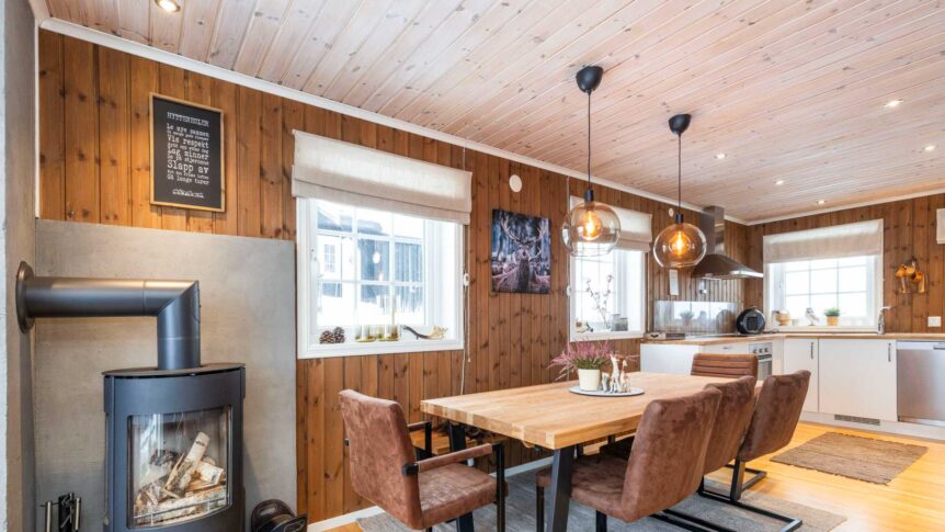 Koselig stue og kjøkken i hyttestil med vedovn, spisebord i tre, og moderne kjøkkeninnredning i naturlig lys.
