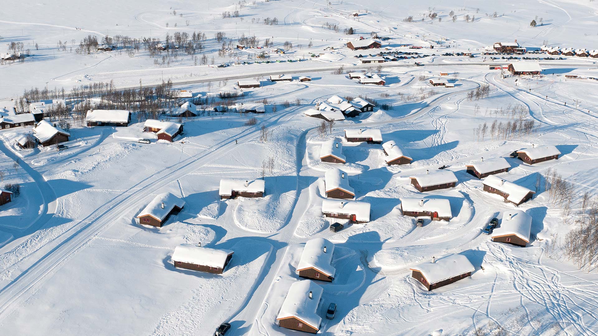Snødekte hytter i et vinterlandskap sett ovenfra, med spor i snøen og skog i bakgrunnen.