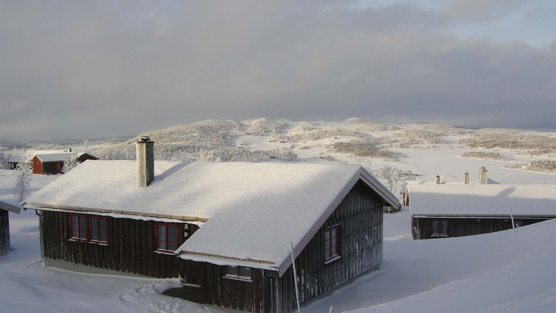 Snødekte norske hytter i vinterlandskap med fjell i bakgrunnen, under grå himmel.