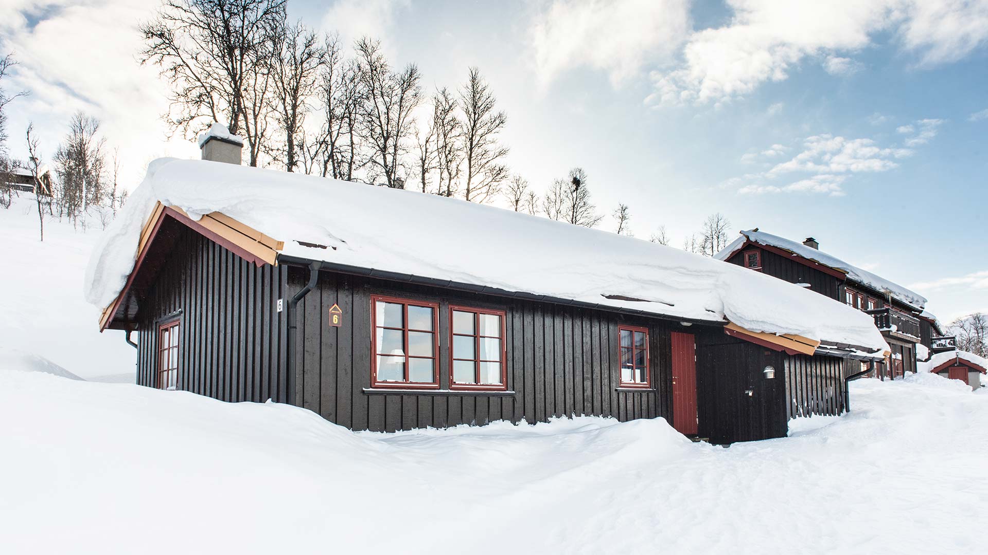 Mørk trehytte i vinterlandskap, dekket av snø, med klare blå himmel bakgrunnen.