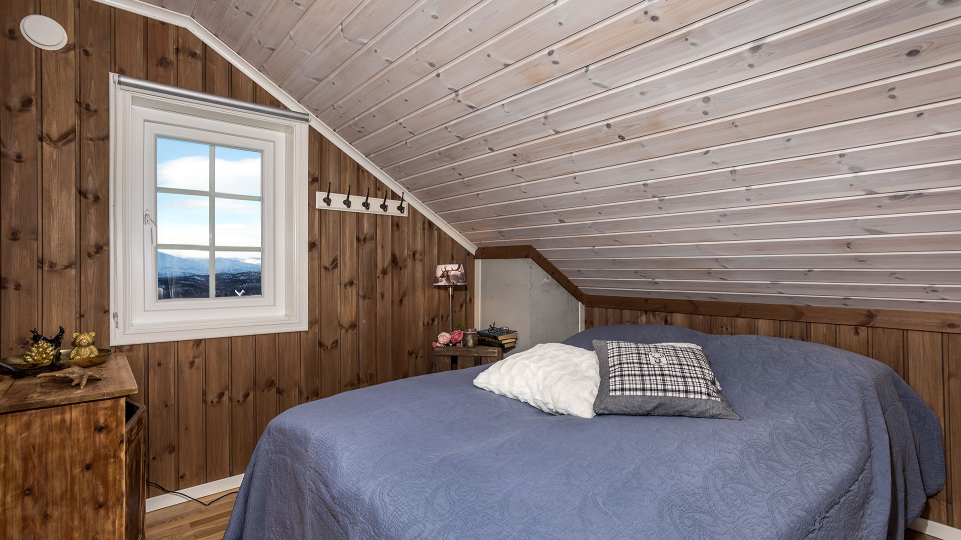 Soverom i loftetasje med trepanel, seng, blått sengeteppe og utsikt mot fjell gjennom vinduet.