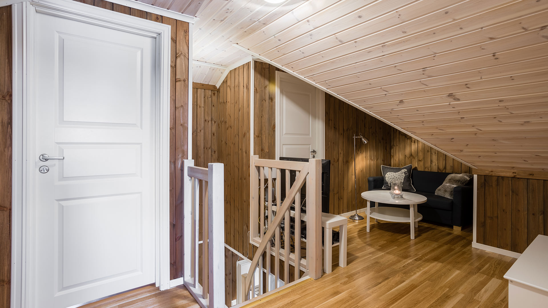 Loftstue med panelvegger i tre, hvit dør, sofa og rundt bord i et koselig, skråtaket interiør.