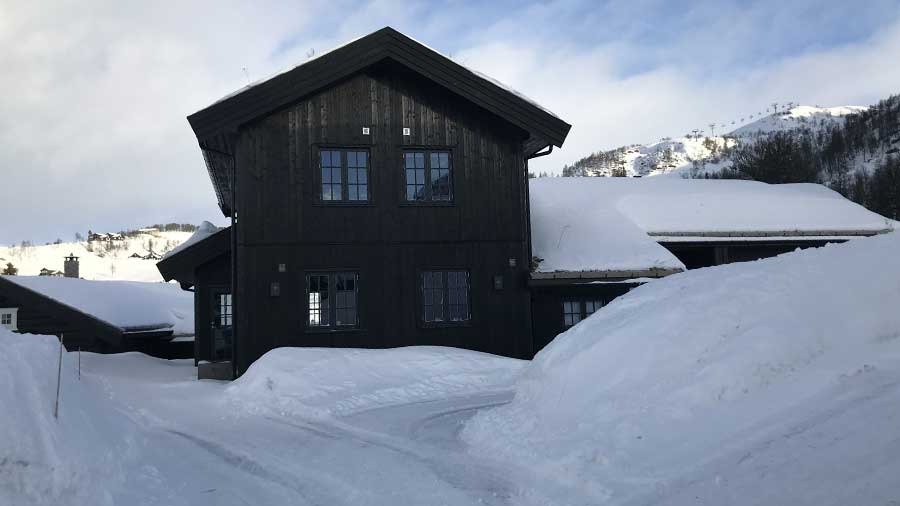 Stor svart hytte i snødekte omgivelser med fjell i bakgrunnen, perfekt for vinterferie i Norge.
