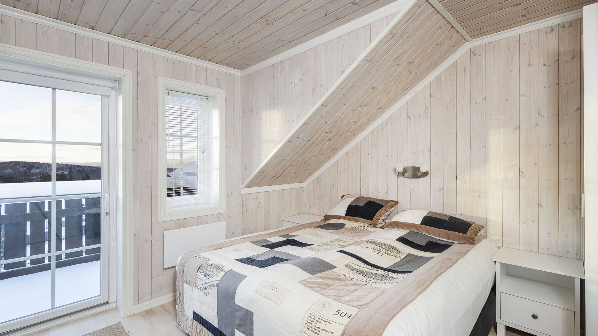 Lyst soverom med dobbeltseng, furupanel og utsikt over snødekt landskap gjennom store vinduer.